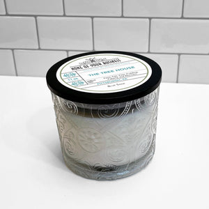 Clear Marquis glass jar with flourish pattern, white soy wax candle. Black lid with label for The Tree House candle. 