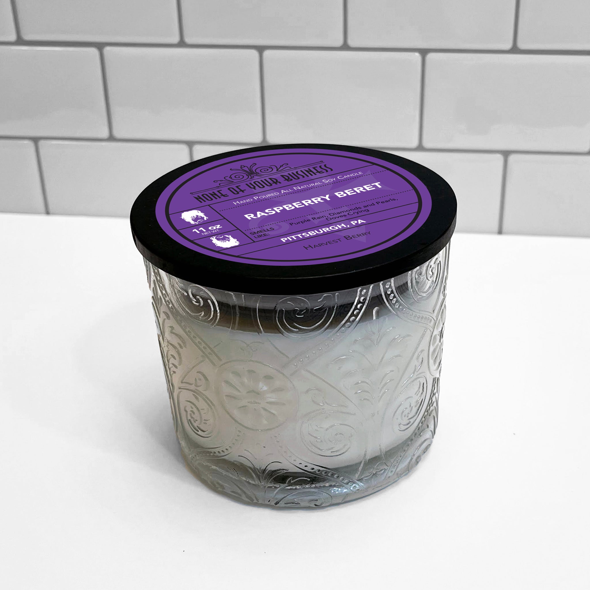 Clear Marquis glass jar with flourish pattern, white soy wax candle. Black lid with label for Raspberry Beret candle. 